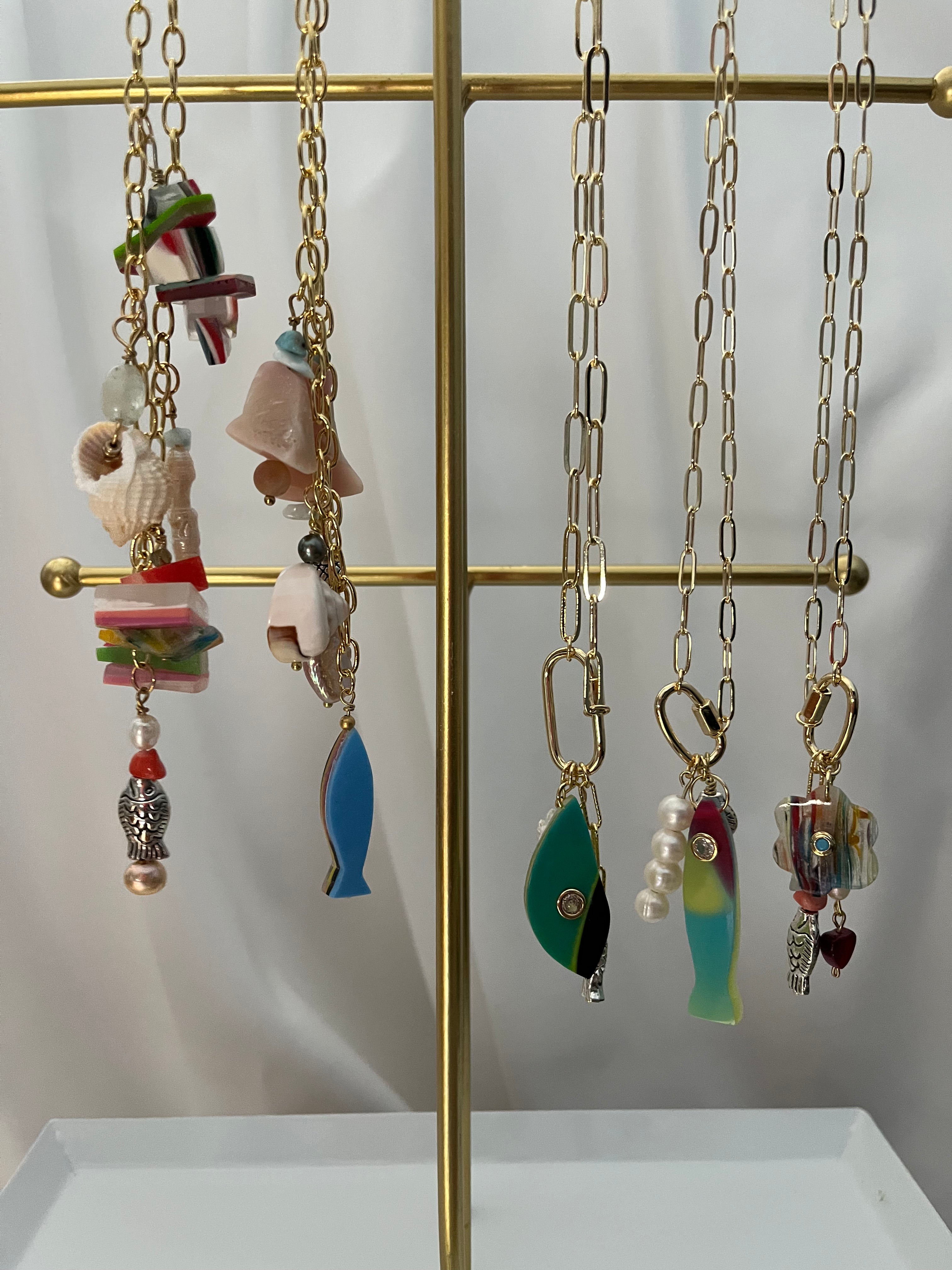 A set of colorful fishing-themed necklaces with various charms (fish, seashells, hooks, beads) hanging from gold-toned chains on a gold display stand.